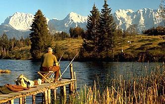 An anterior man sitting on a foortbridge while fishing in the lake, in the background the Alps